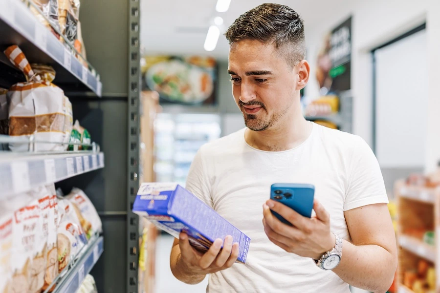 A young Caucasian man is holding his cellphone and checking the ingredients on a box of breadsticks while standing in a supermarket.